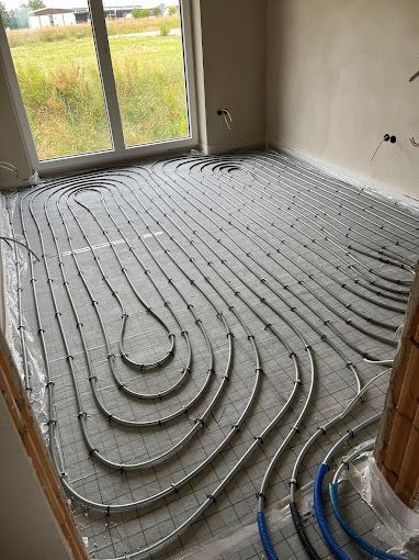 In einem Raum mit Betonwänden und einem großen Fenster sind die Rohre einer Fußbodenheizung gitterförmig verlegt.