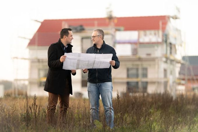 Zwei Männer stehen auf einem Feld und betrachten einen Bauplan vor einem im Bau befindlichen Haus.