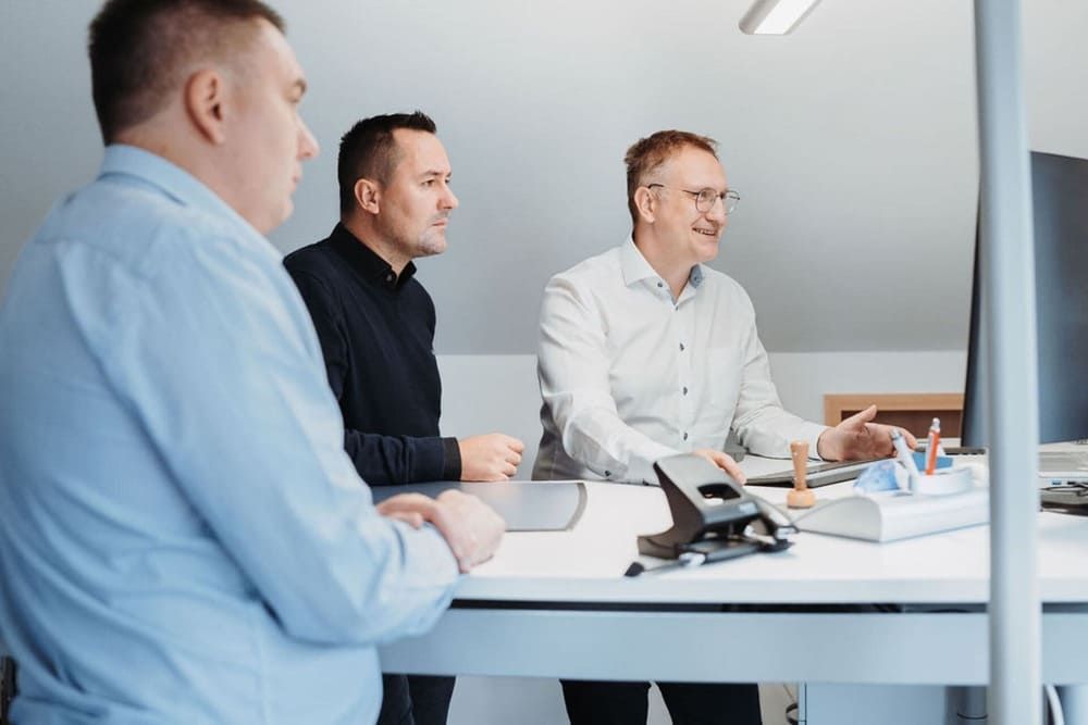 Drei Männer sitzen an einem Schreibtisch vor einem Computer.