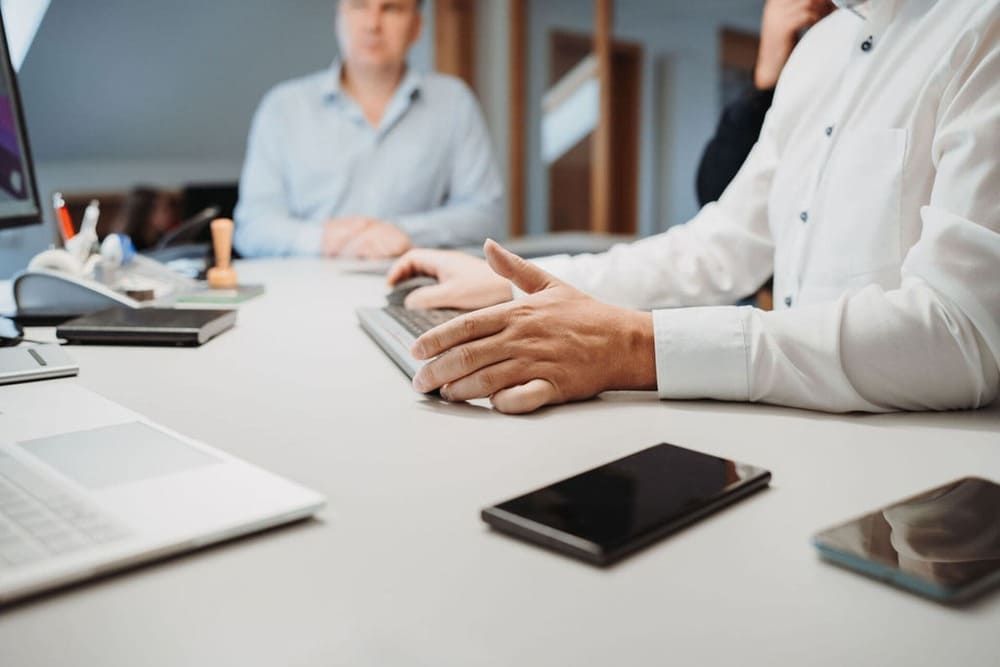 Ein Mann sitzt mit einem Laptop und einem Handy an einem Schreibtisch.