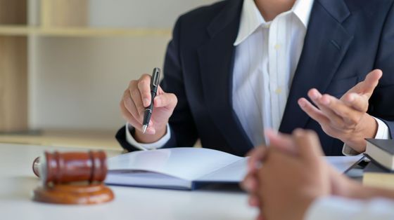 Un abogado con traje gesticula mientras habla con un cliente en un escritorio, con el mazo en primer plano.