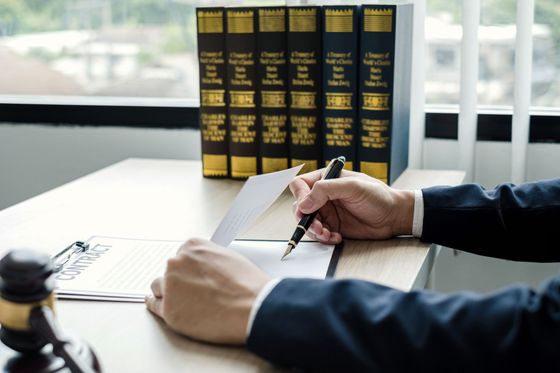 Una persona con traje firma un documento sobre un escritorio, con libros de leyes y un mazo visibles.