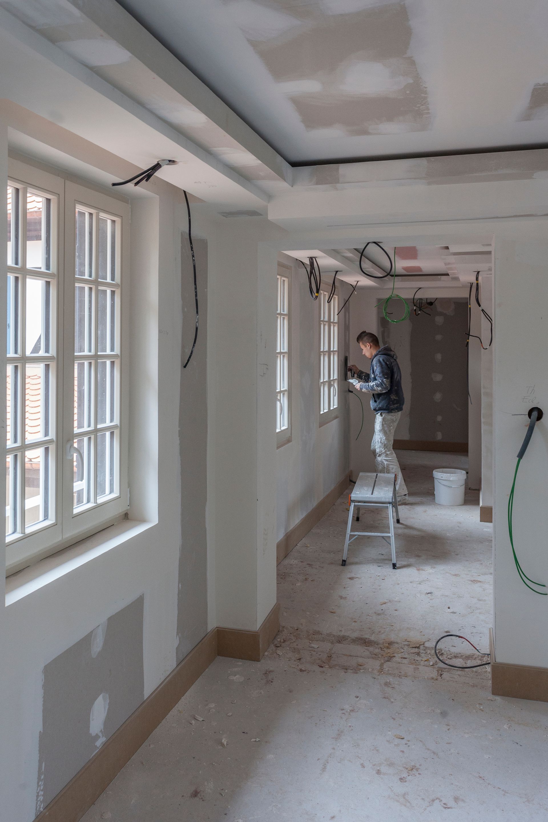 Ouvrier du bâtiment sur un escabeau travaillant sur le câblage électrique dans une pièce avec des fenêtres et des fils exposés.