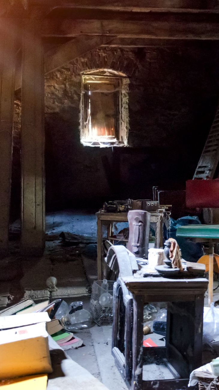 Grenier poussiéreux, baigné de soleil par une fenêtre. Tables de travail et débris éparpillés ; éclairage tamisé.