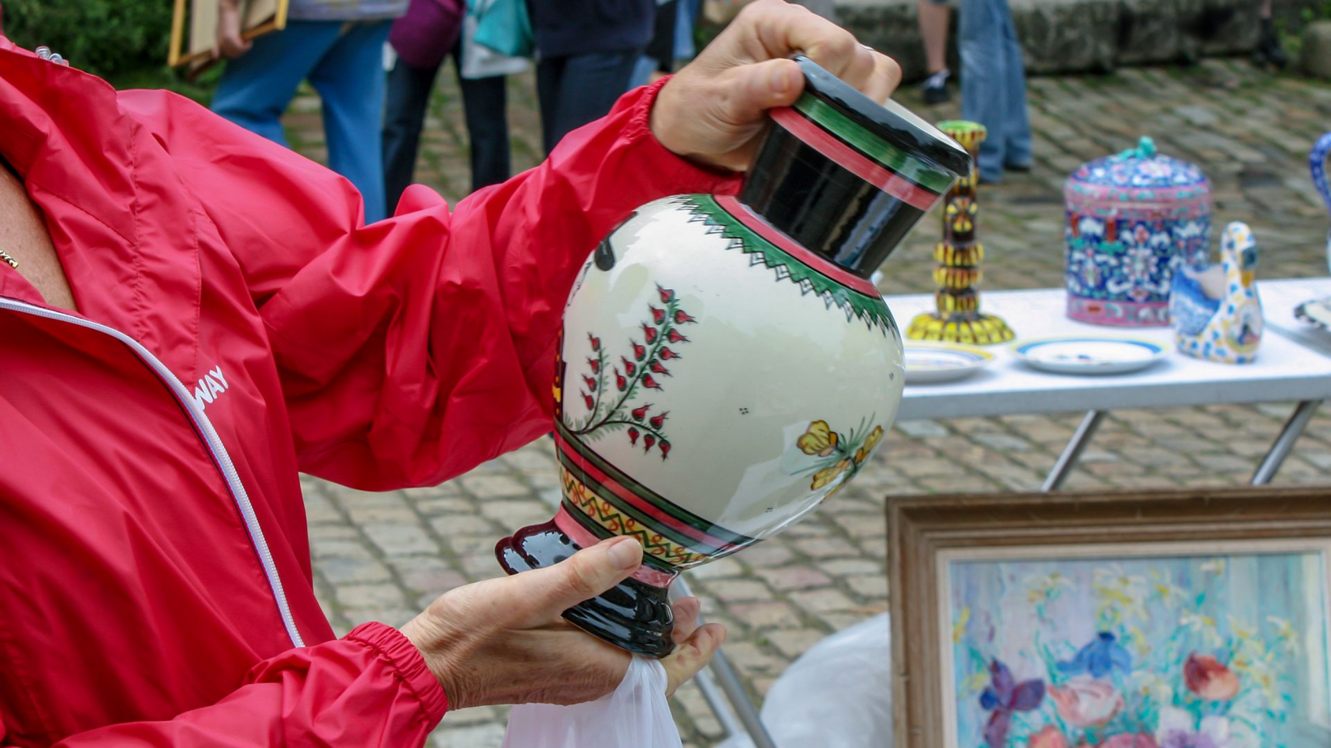 Personne en veste rouge tenant un vase décoratif blanc et noir portant des détails de feuillage sur un marché en plein air.