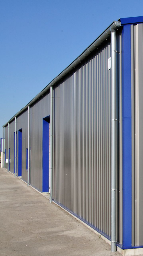 Long bâtiment industriel avec revêtement en tôle ondulée, cadres de portes bleus et chemin en béton.