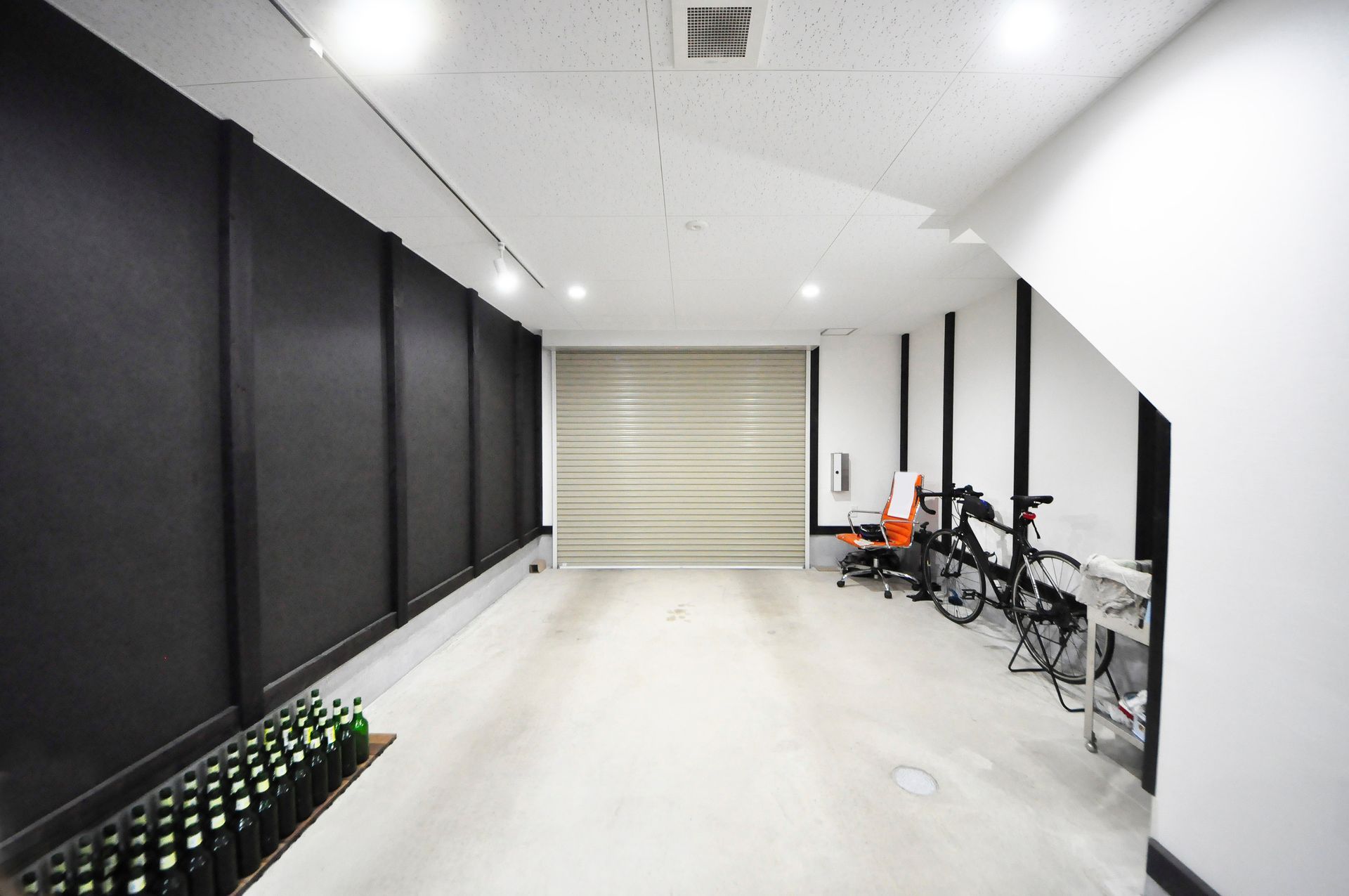 Pièce vide avec rideaux sombres à gauche et porte de garage fermée. Vélo et chaise de bureau visibles.