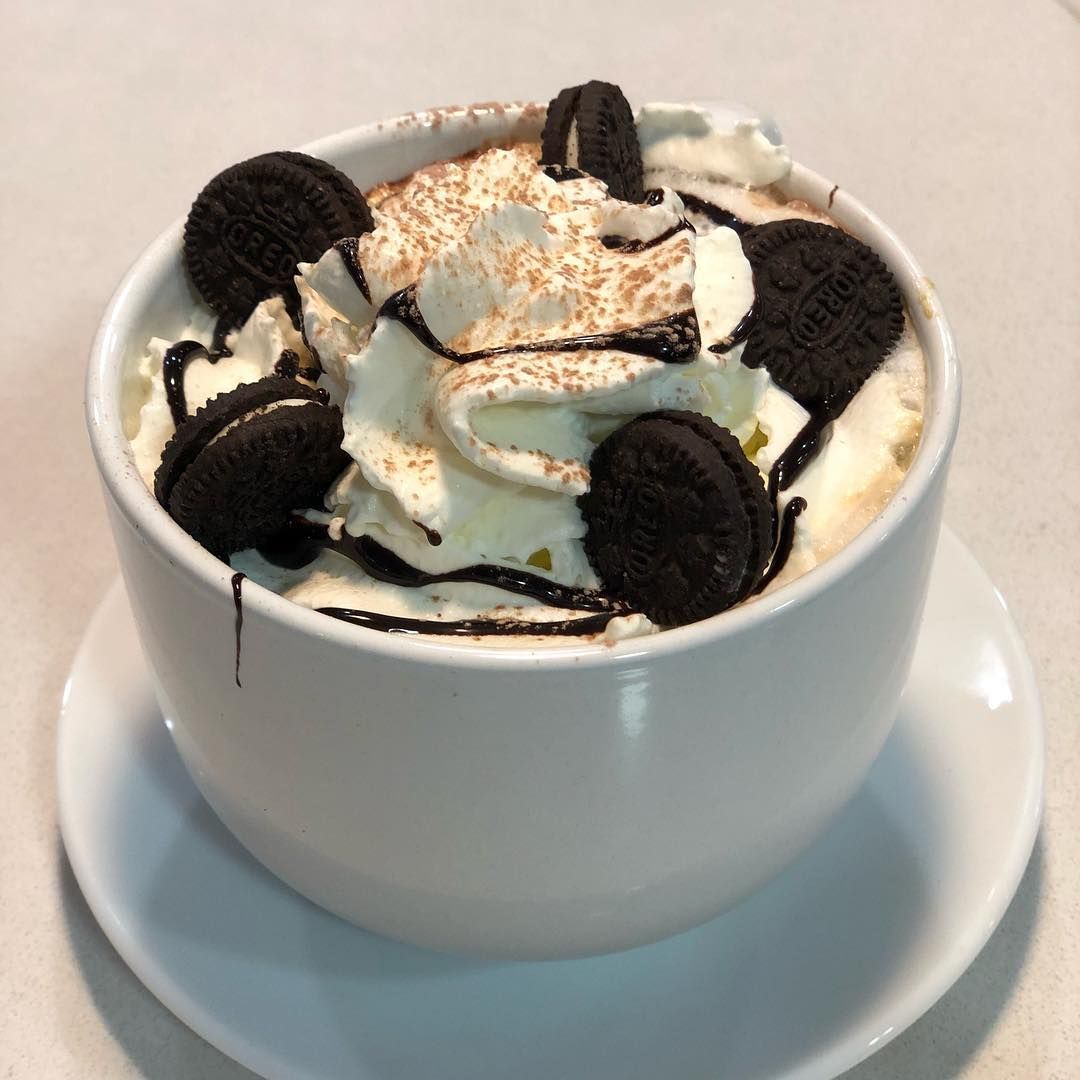 Una taza blanca llena de una bebida de postre, cubierta con crema batida, sirope de chocolate y mini galletas tipo sándwich.