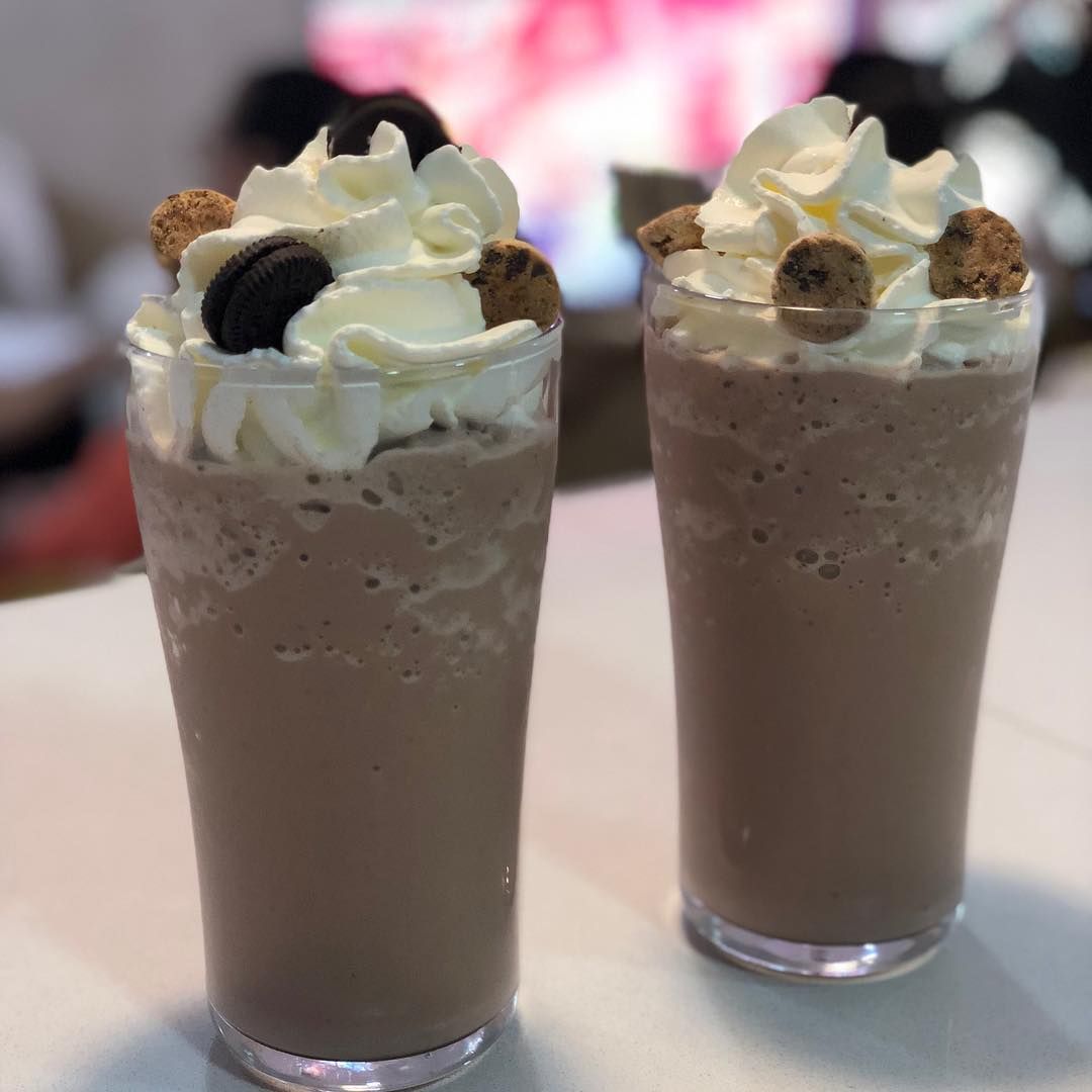 Dos vasos altos de batido de chocolate cubiertos con crema batida, galletas de chocolate rellenas y trocitos de galleta.