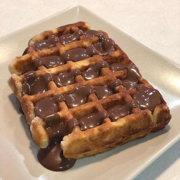 Un gofre cuadrado cubierto con salsa de chocolate derretido reposa sobre un plato blanco.