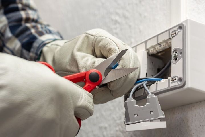 Eine Person mit Schutzhandschuhen benutzt eine rote Schere, um blaue elektrische Leitungen abzuschneiden, die an eine Wandsteckdose angeschlossen sind.