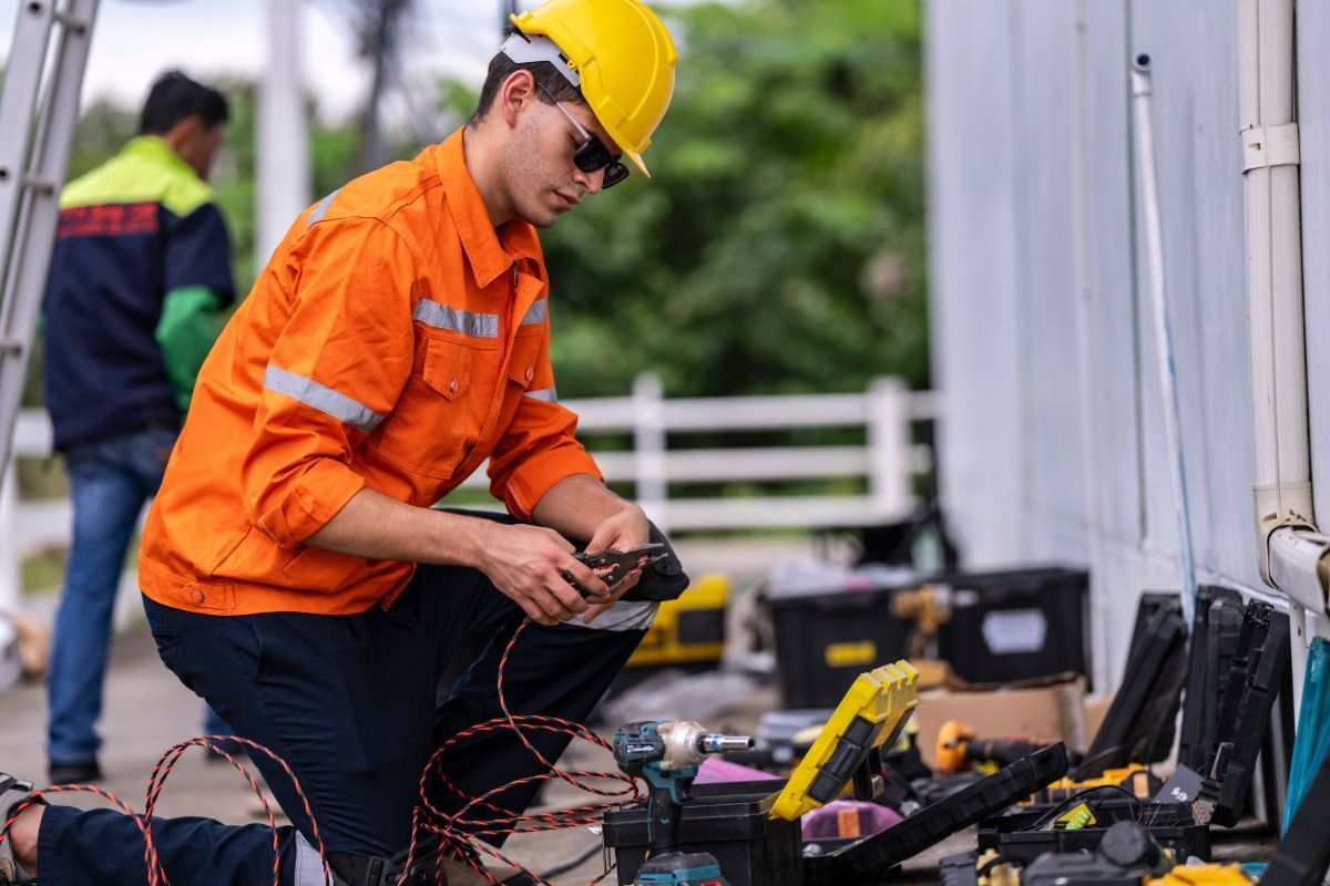 Ein Arbeiter in einer gut sichtbaren orangefarbenen Jacke und einem gelben Schutzhelm kniet, um elektrische Leitungen auf einer Baustelle zu überprüfen.