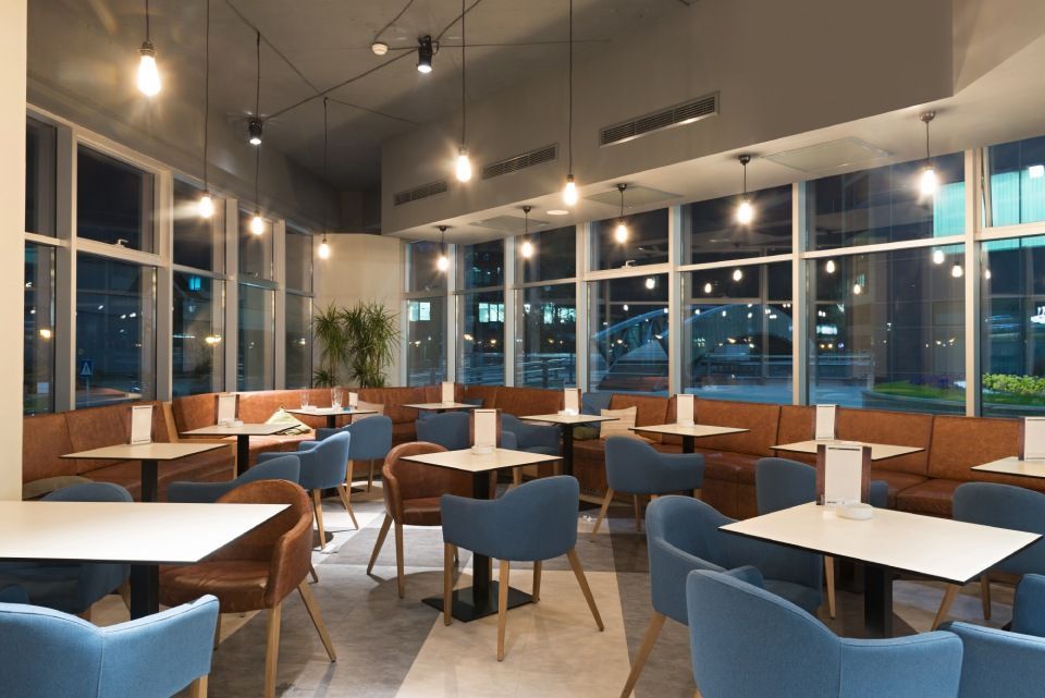 Das Interieur eines modernen Restaurants bei Nacht zeigt braune Sitznischen, weiße Tische und blaue Stühle unter warmen Hängelampen.