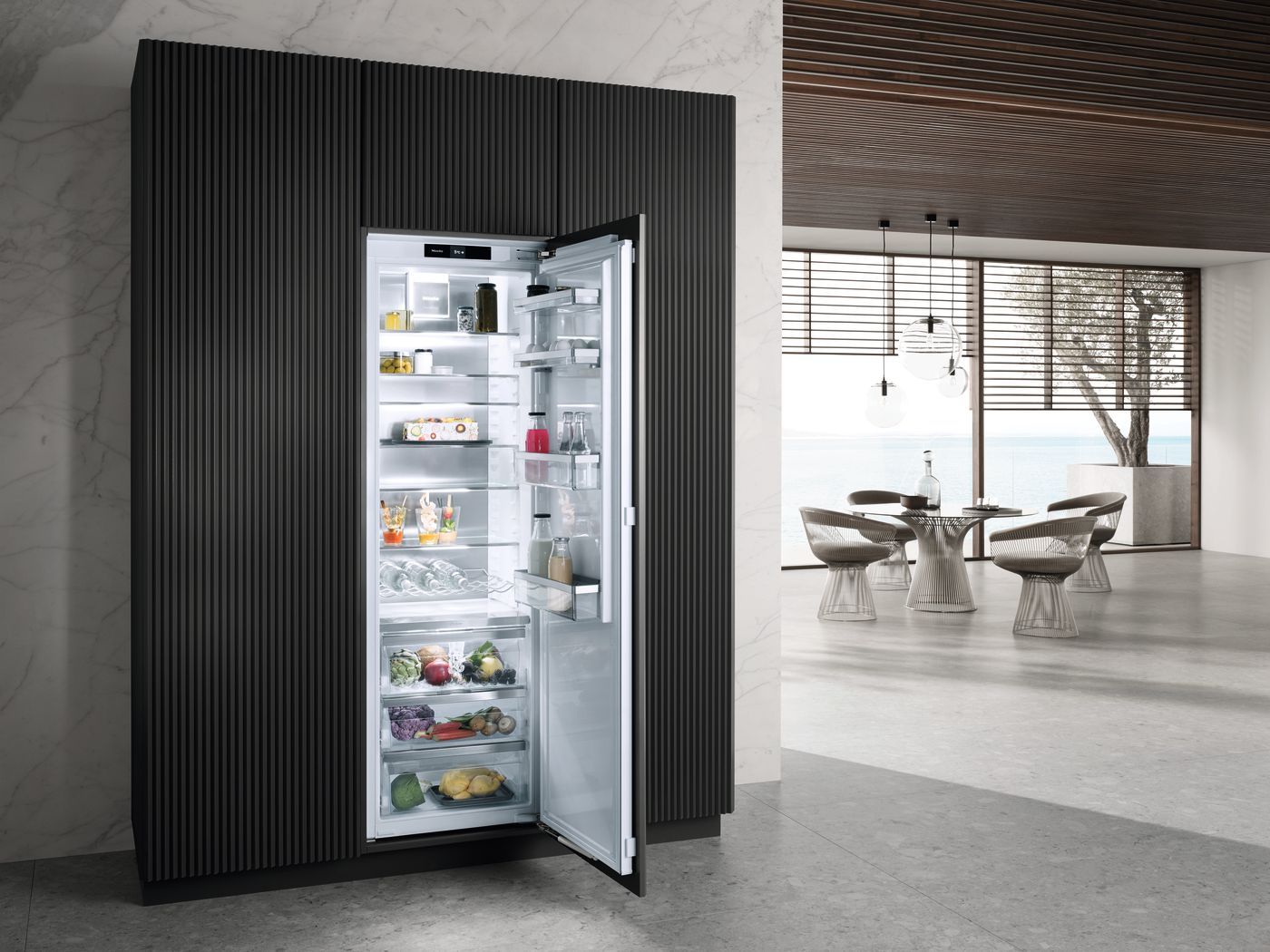 Ein offener, integrierter Kühlschrank, der in eine dunkle, strukturierte Paneelwand in einem modernen Raum mit großen Fenstern eingebaut ist.