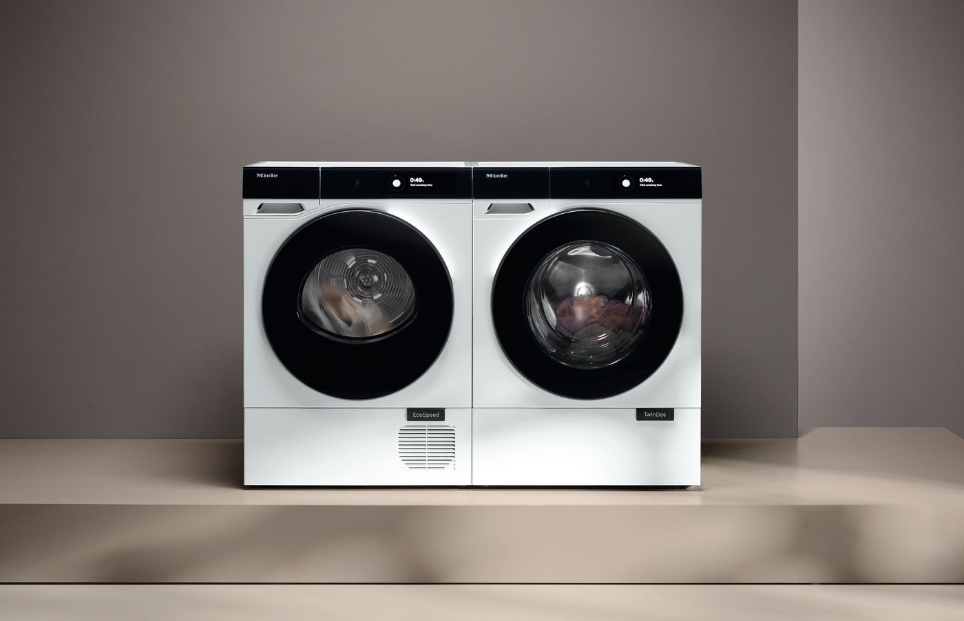 In einem grauen Waschraum steht eine weiße Waschmaschine zwischen einem Metallregal mit Handtüchern und einem Stuhl mit Kissen.