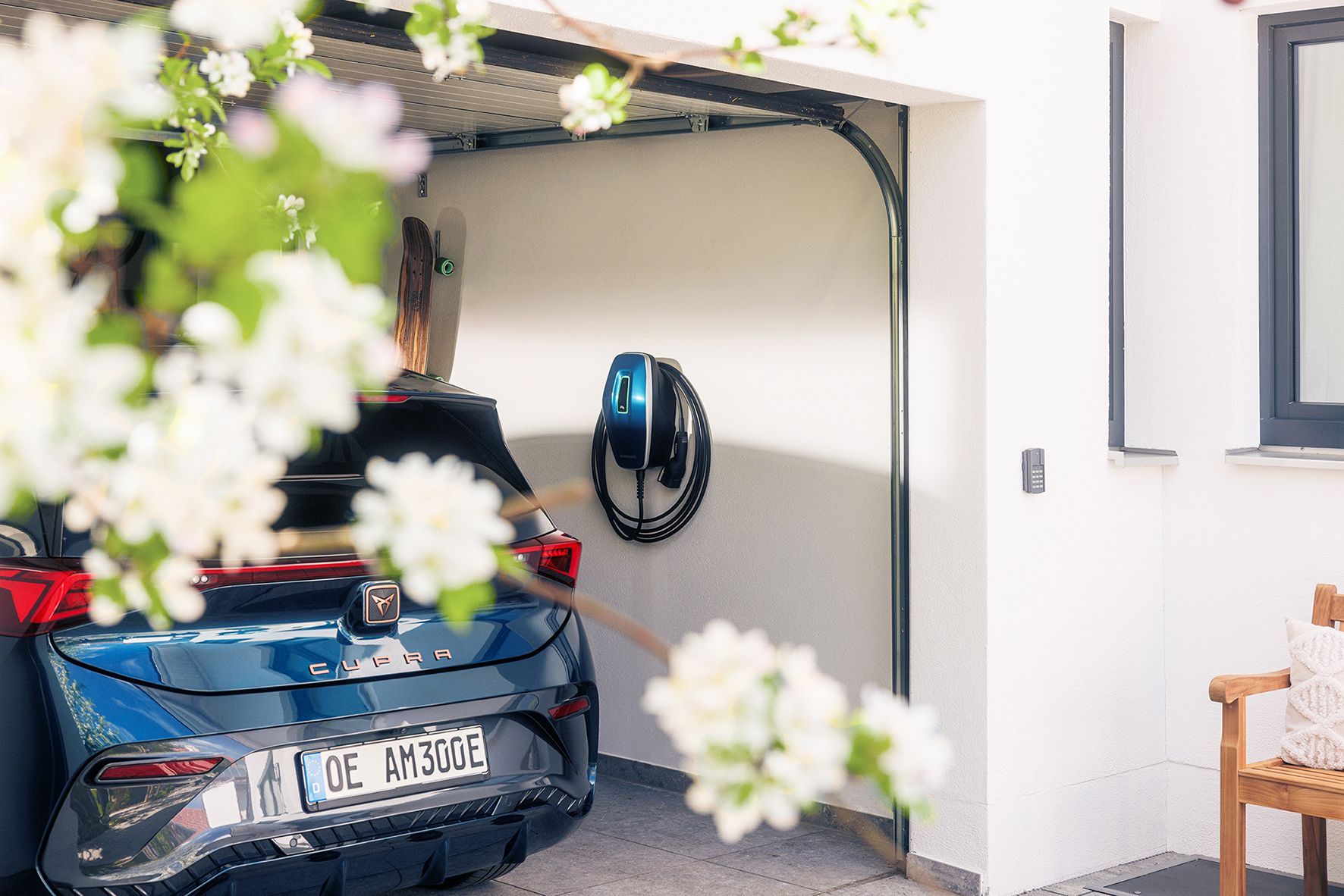 Ein dunkelblaues Elektroauto, geparkt in einer Garage mit einer an der Wand montierten Ladestation, umrahmt von weißen Frühlingsblumen.