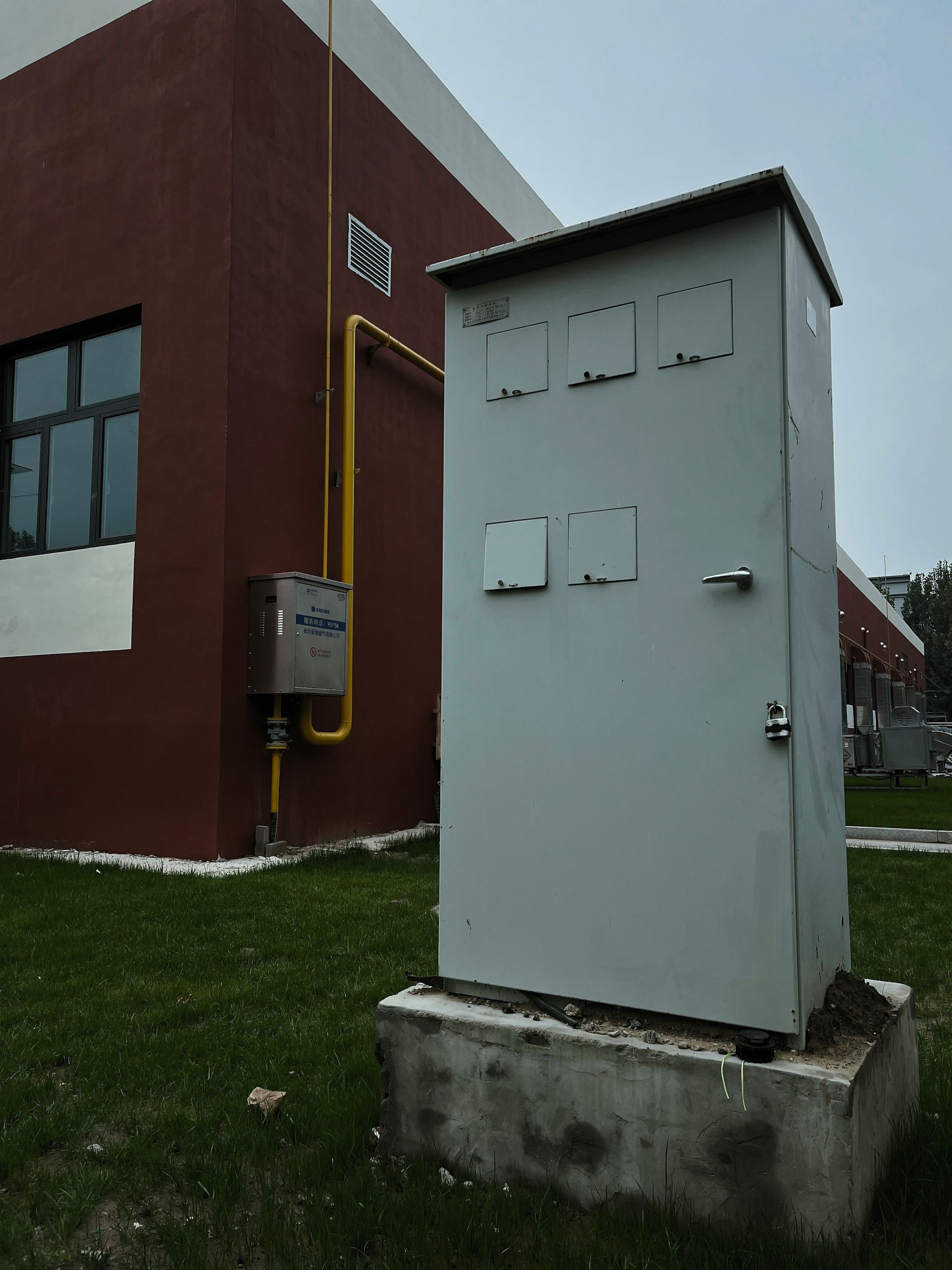 Ein hoher, hellgrauer Geräteschrank steht auf einem Betonsockel neben einem kastanienbraunen Gebäude mit gelben Gasleitungen.