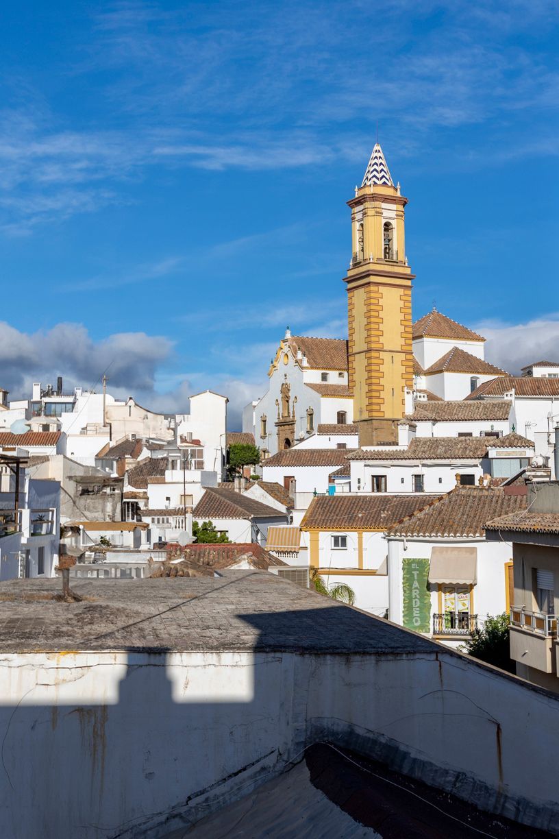 Vista de edificios blancos con techos de tejas naranjas y una alta torre de iglesia amarilla contra un cielo azul.