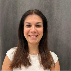 Mujer con cabello largo y castaño, sonriente, vistiendo una camisa blanca, sobre un fondo gris.