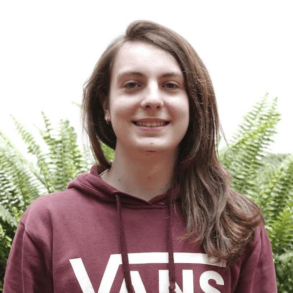 Joven de pelo castaño corto, sonriente, con chaqueta negra sobre camisa a rayas. Frente a una pared beige.
