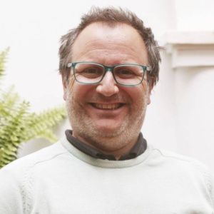 Un hombre con gafas sonríe, vistiendo un suéter verde claro, al aire libre frente a una pared blanca con una planta.