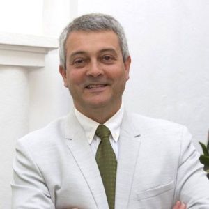 Hombre de mediana edad, de cabello gris, sonriente, vestido con traje blanco y corbata verde, con los brazos cruzados.