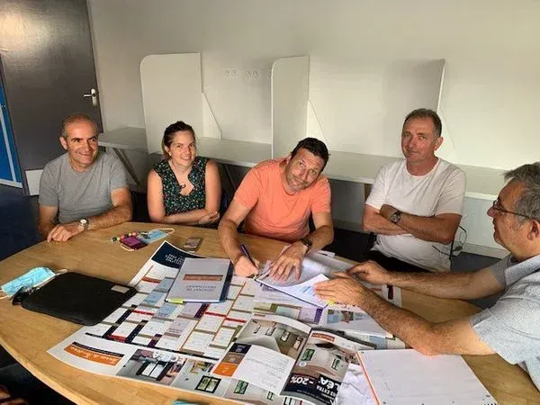 Cinq personnes assises autour d'une table, examinant des documents et les signant. Intérieur lumineux.