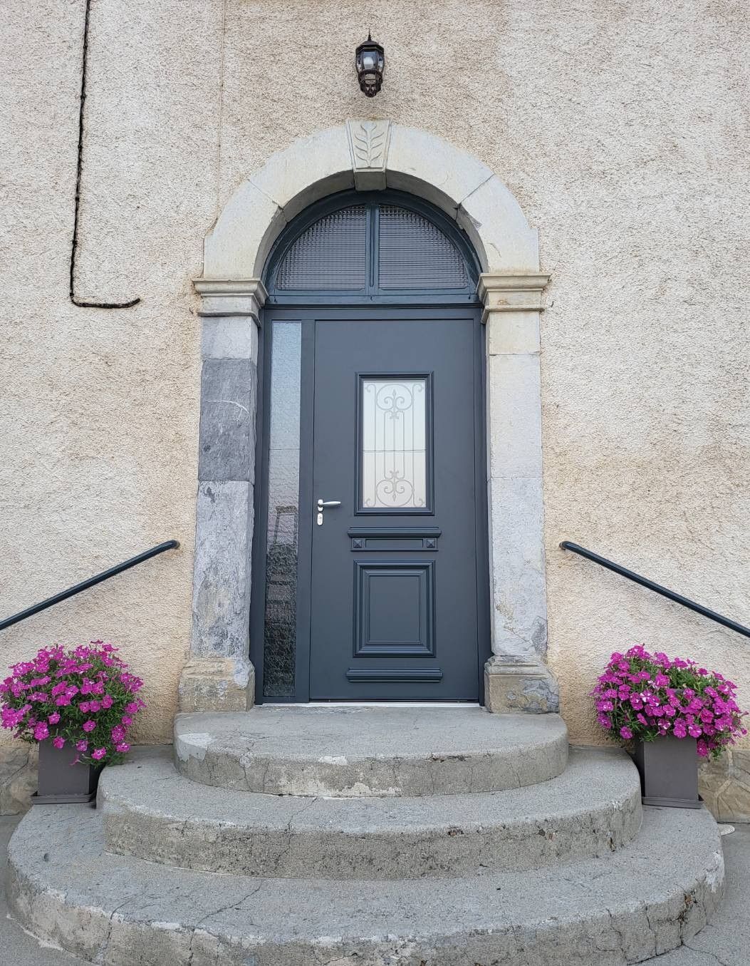 Porte d'entrée blanche avec fenêtres latérales, flanquée de plantes en pot et d'appliques sur une maison de couleur claire.