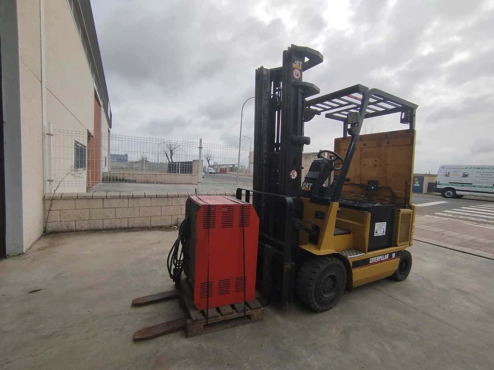 Una carretilla elevadora eléctrica amarilla está estacionada al aire libre en un terreno de hormigón, con un cargador de baterías de metal rojo sujeto a sus horquillas.