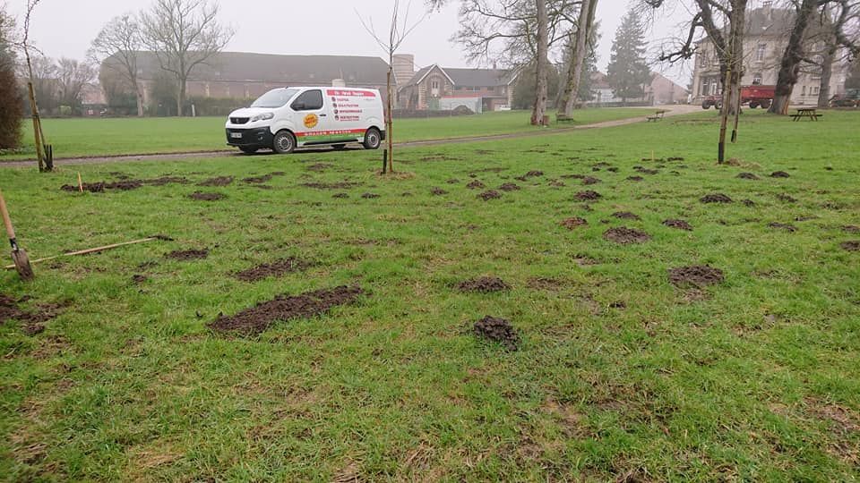 Une camionnette blanche roule sur un champ herbeux parsemé de monticules de terre, avec des bâtiments en arrière-plan.