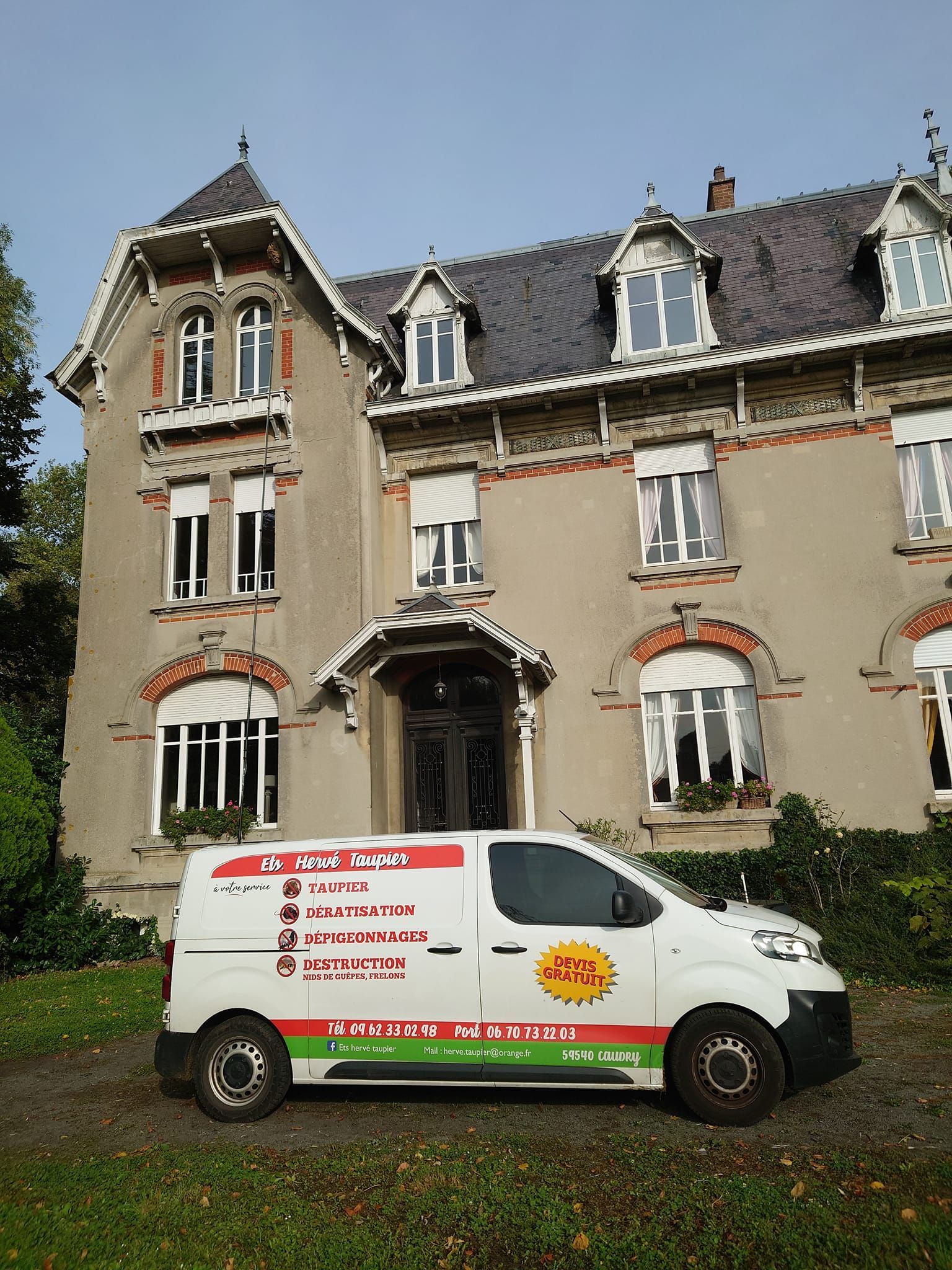 Une camionnette blanche avec un logo est garée devant une grande maison grise à plusieurs fenêtres.