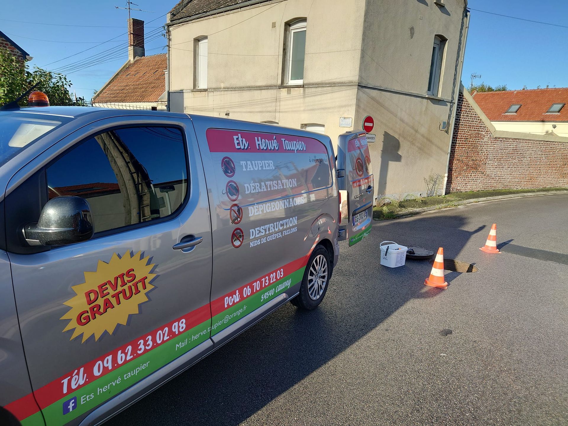 Fourgonnette grise garée dans une rue bordée de cônes orange. Bâtiment en arrière-plan.