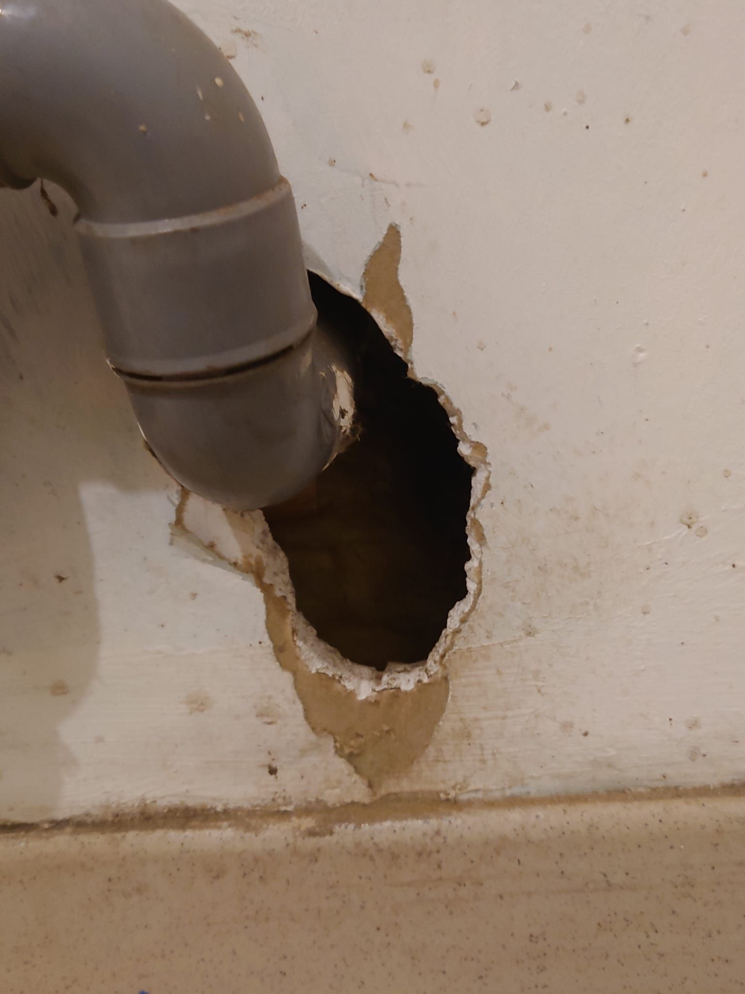 Un tuyau gris dépasse d'un grand trou dans un mur blanc ; des débris entourent le trou.