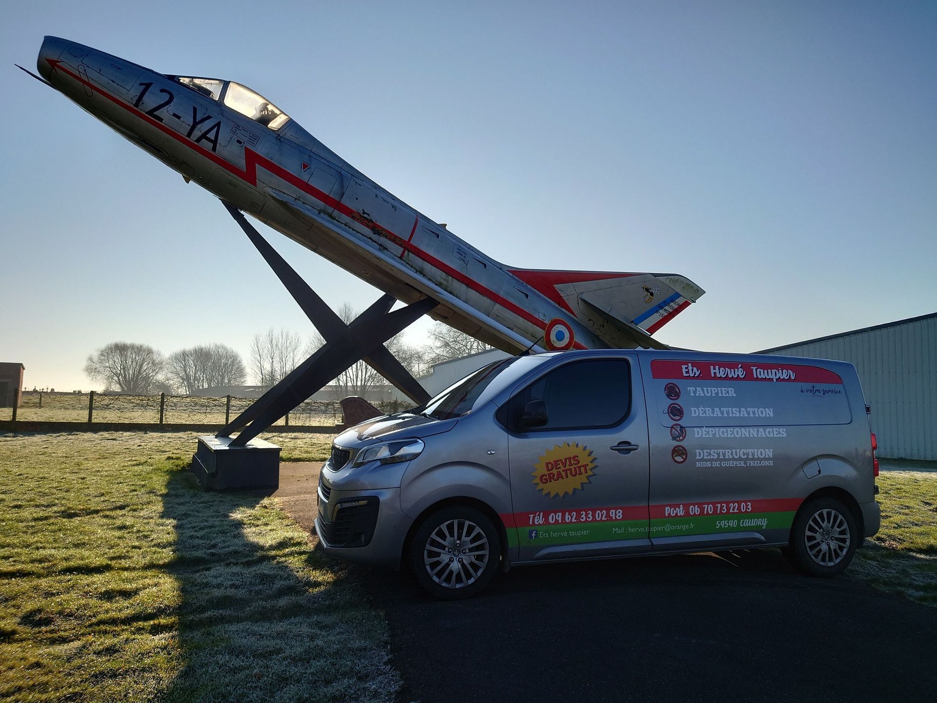 Une camionnette grise arborant le logo de l'entreprise est garée devant un avion à réaction argenté stationné lui aussi. Journée ensoleillée.