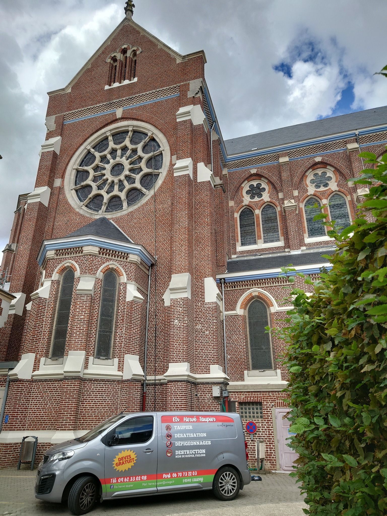 Une camionnette garée devant une église en briques ornée d'une grande rosace et d'une architecture décorative, sous un ciel nuageux.