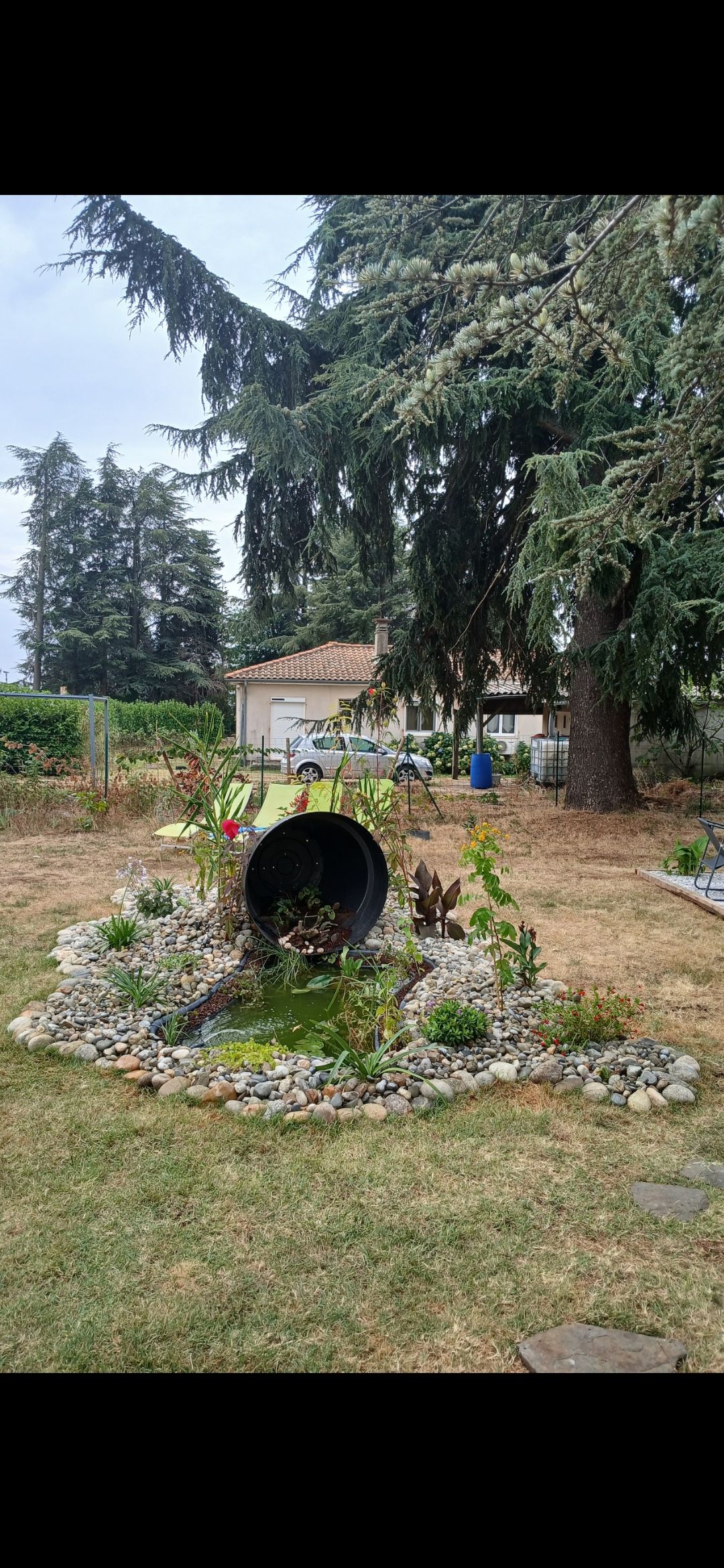 Une scène de jardin avec une grande canalisation entourée de pierres et de verdure ; une maison se trouve en arrière-plan.