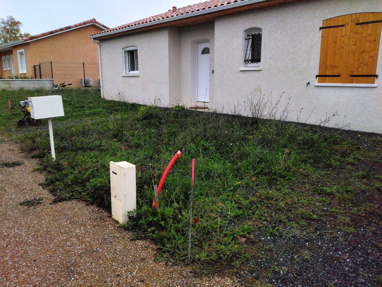Maison avec pelouse non entretenue, boîte aux lettres et boîtiers électriques. Des tuyaux rouges sont visibles.