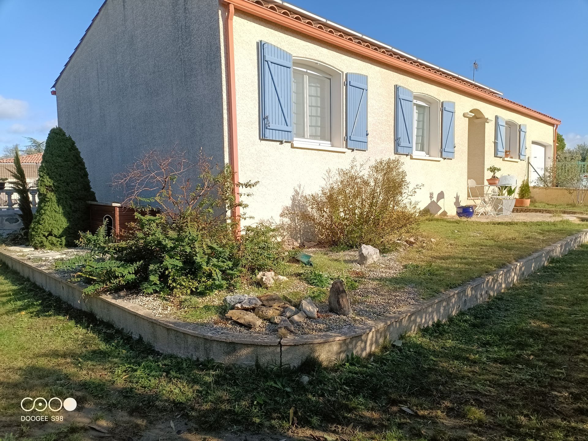 Maison beige avec des volets bleus, petit jardin bordé de pavés.