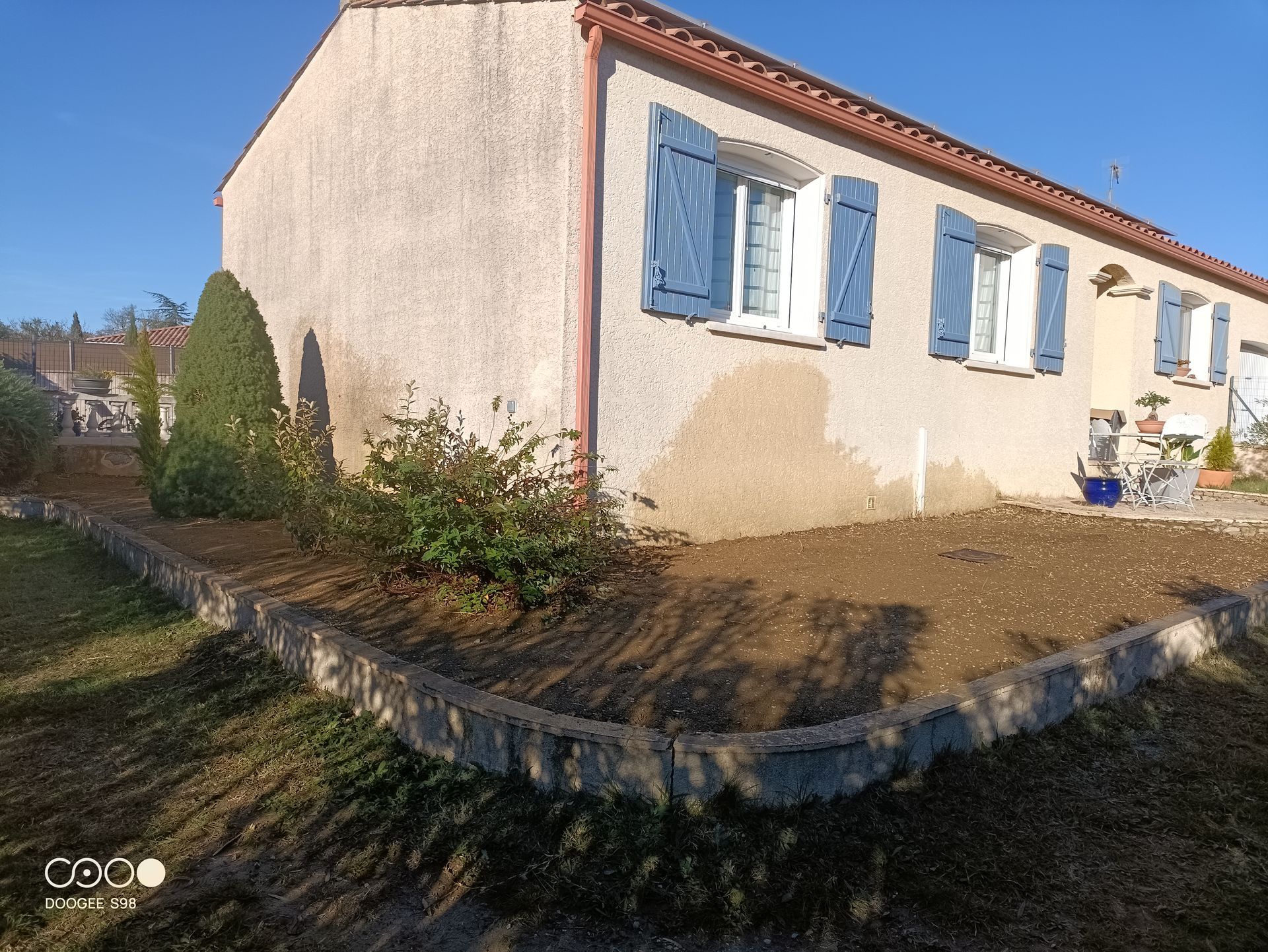 Maison beige avec volets bleus, toit de tuiles rouges et parterre de jardin bas en pierre.