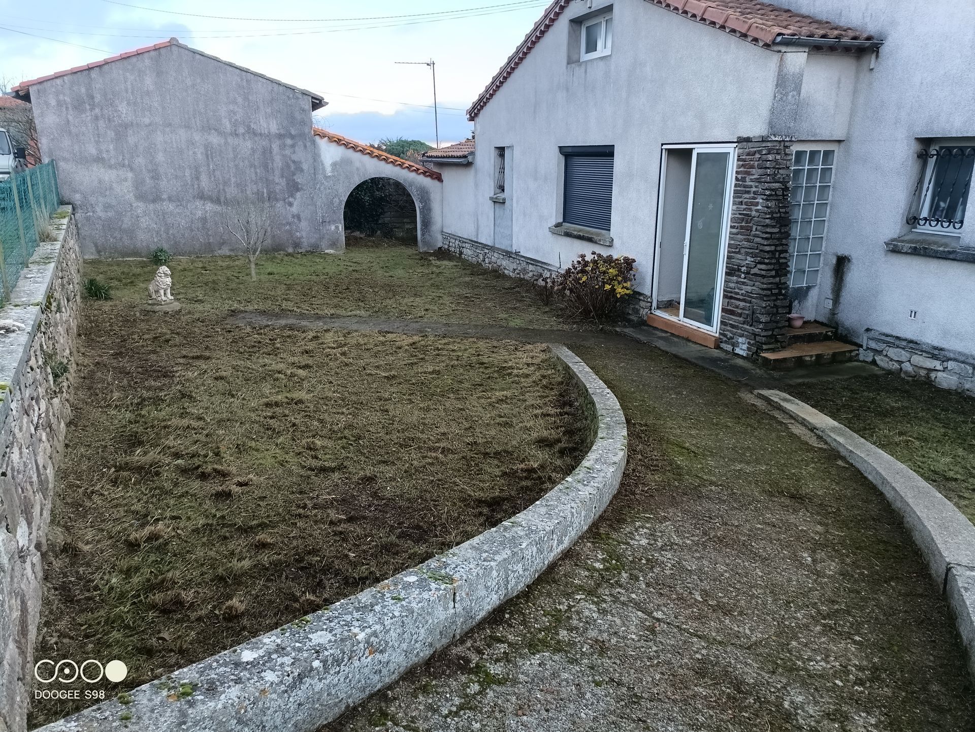Une maison avec un jardin. Un chemin de pierre sinueux mène du premier plan à la maison et à son dépendance. Ciel couvert.