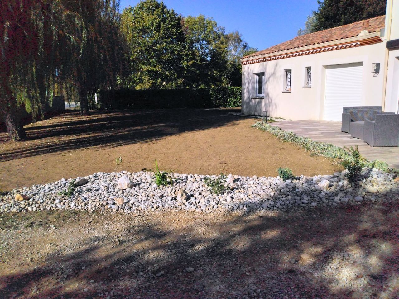 L'extérieur d'une maison avec un lit de gravier, une pelouse fraîchement semée et des arbres sous un ciel bleu limpide.