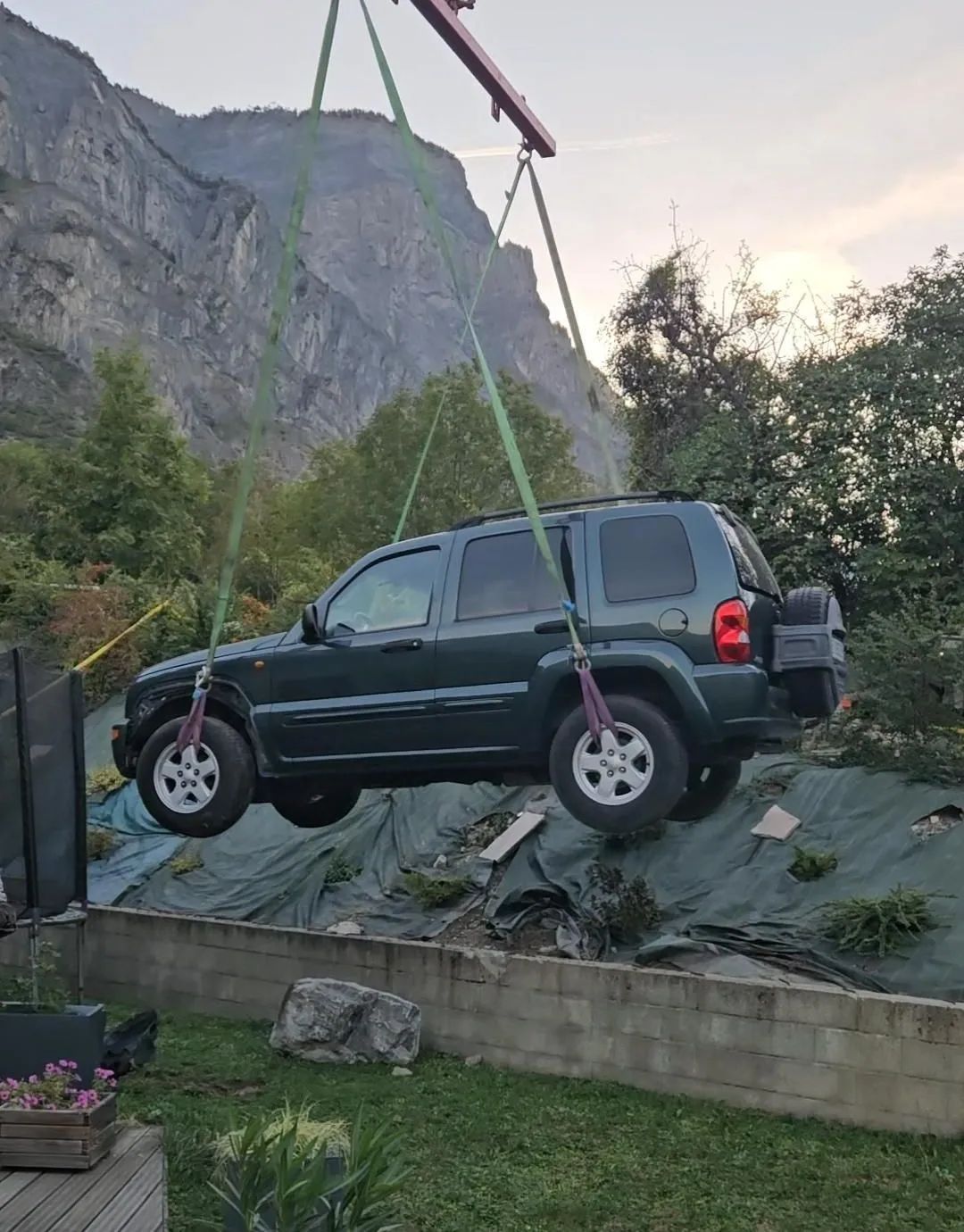 Jeep SUV verte suspendue par une grue ; montagnes et végétation en arrière-plan.