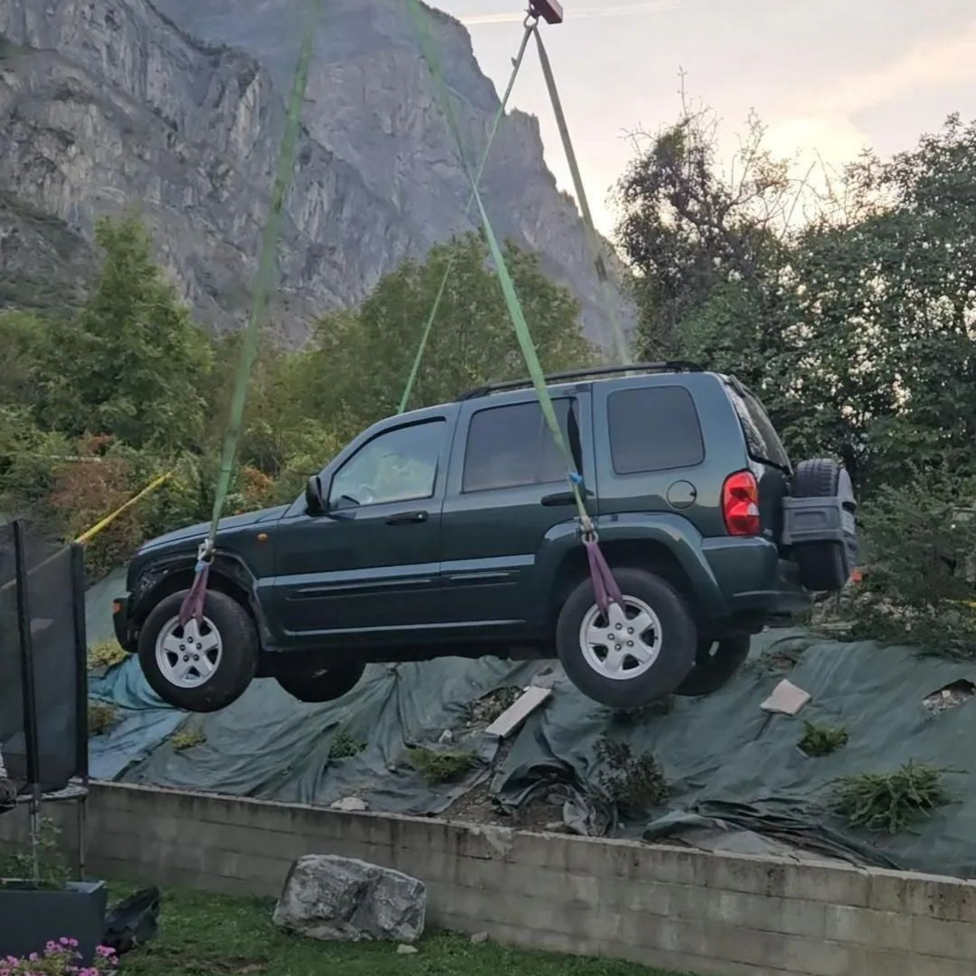 Jeep verte soulevée par une grue, suspendue au-dessus d'une colline, montagnes en arrière-plan.