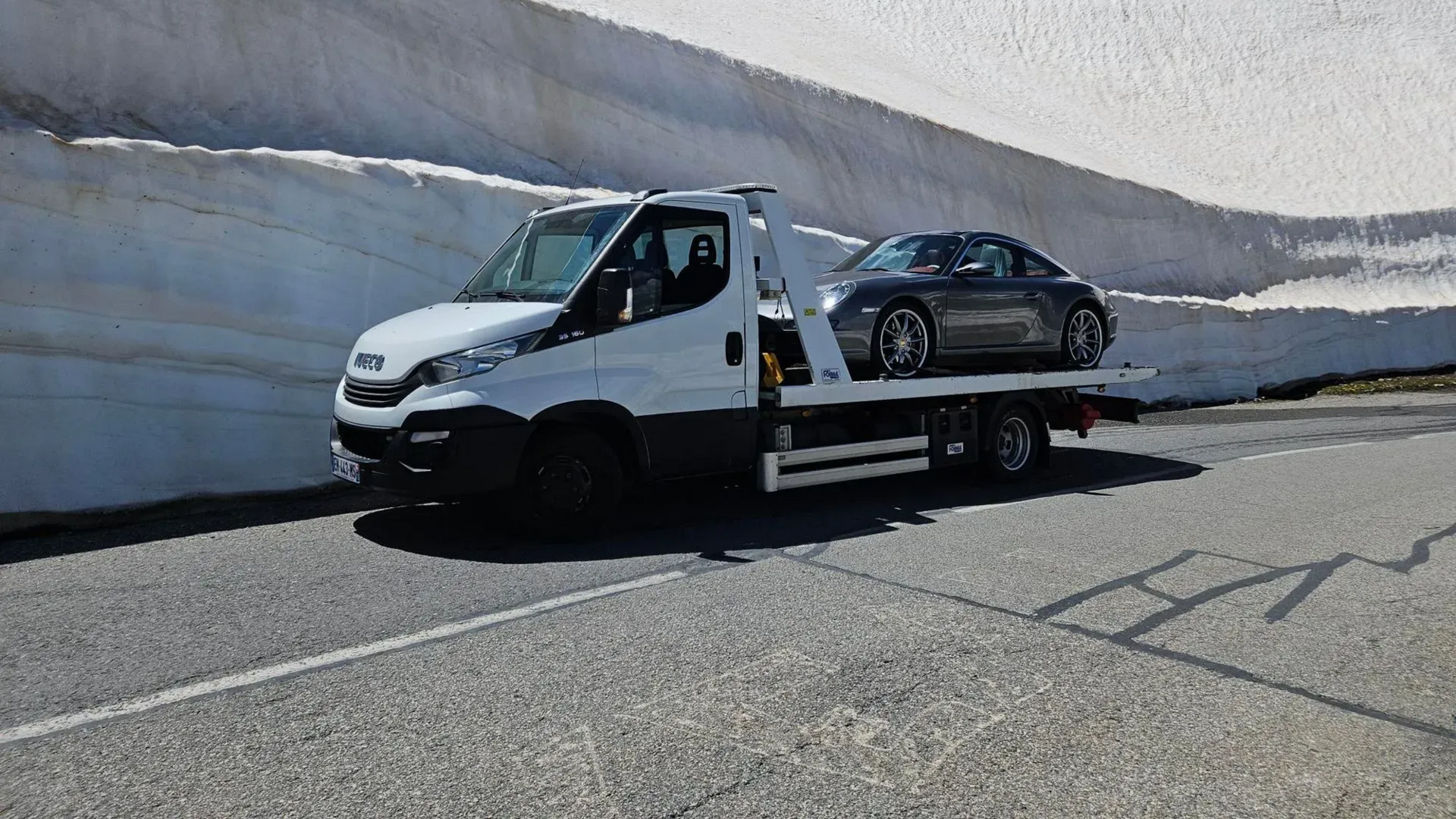 Une dépanneuse transporte une voiture de sport grise sur une route bordée d'un banc de neige.