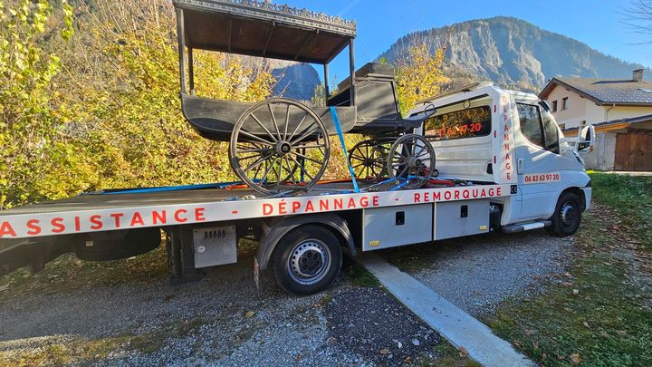 Une dépanneuse transporte une calèche ancienne sur son plateau, garée à l'extérieur près des montagnes et des arbres.