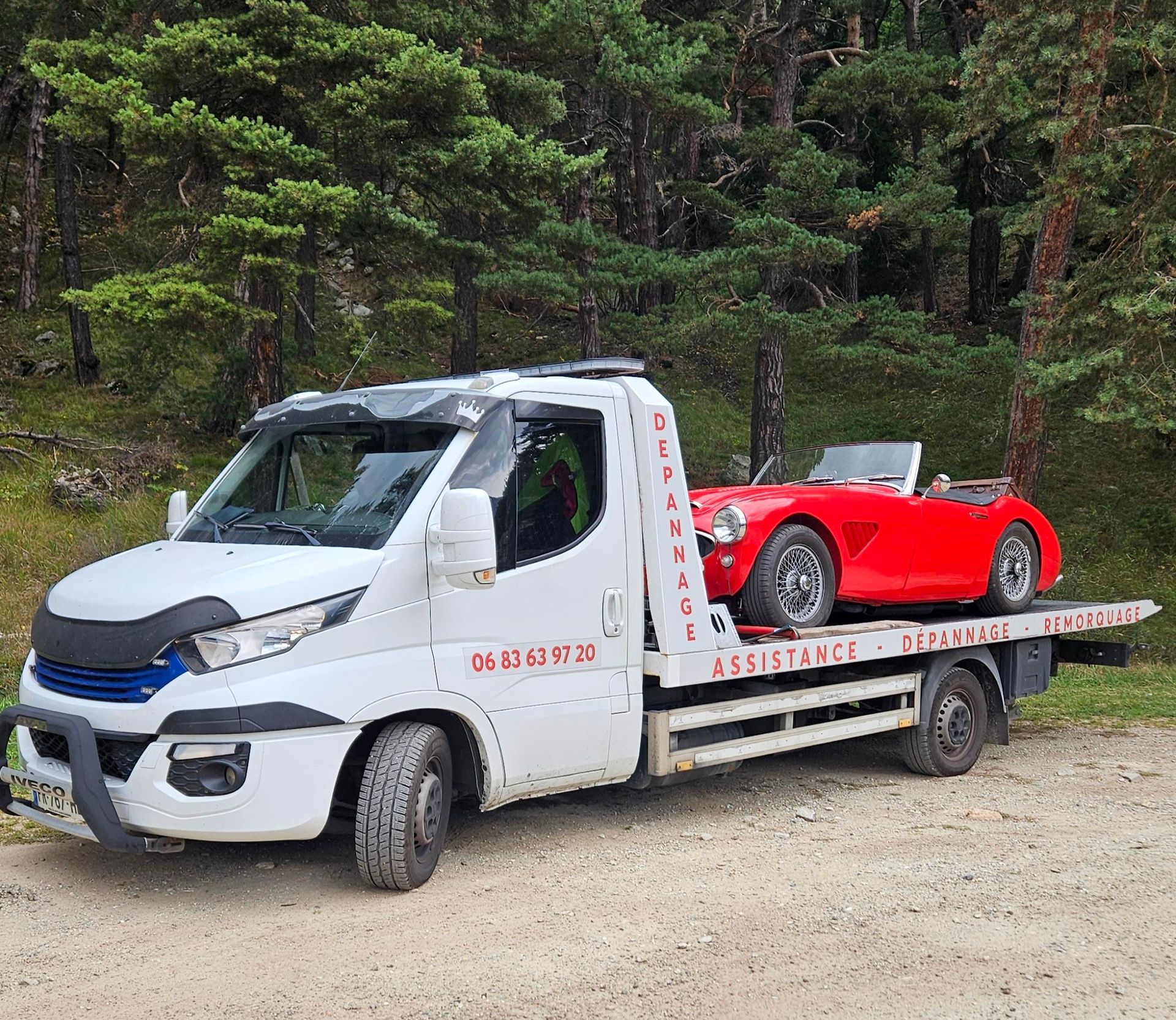 Une dépanneuse blanche transporte une voiture de sport classique rouge sur une route boisée.