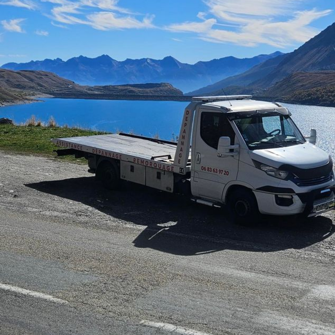 Un camion de dépannage blanc est stationné près d'un lac, avec des montagnes en arrière-plan sous un ciel bleu.