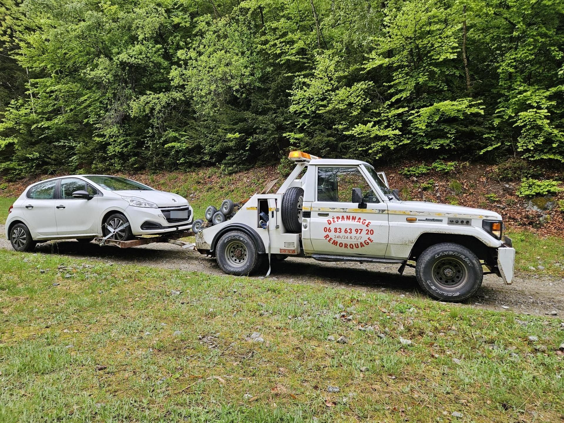Un camion de dépannage blanc tire une voiture blanche sur un chemin de terre, des arbres se dressent en arrière-plan.