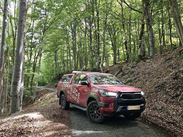 Un pick-up rouge roule sur un chemin de gravier à travers une forêt verdoyante.