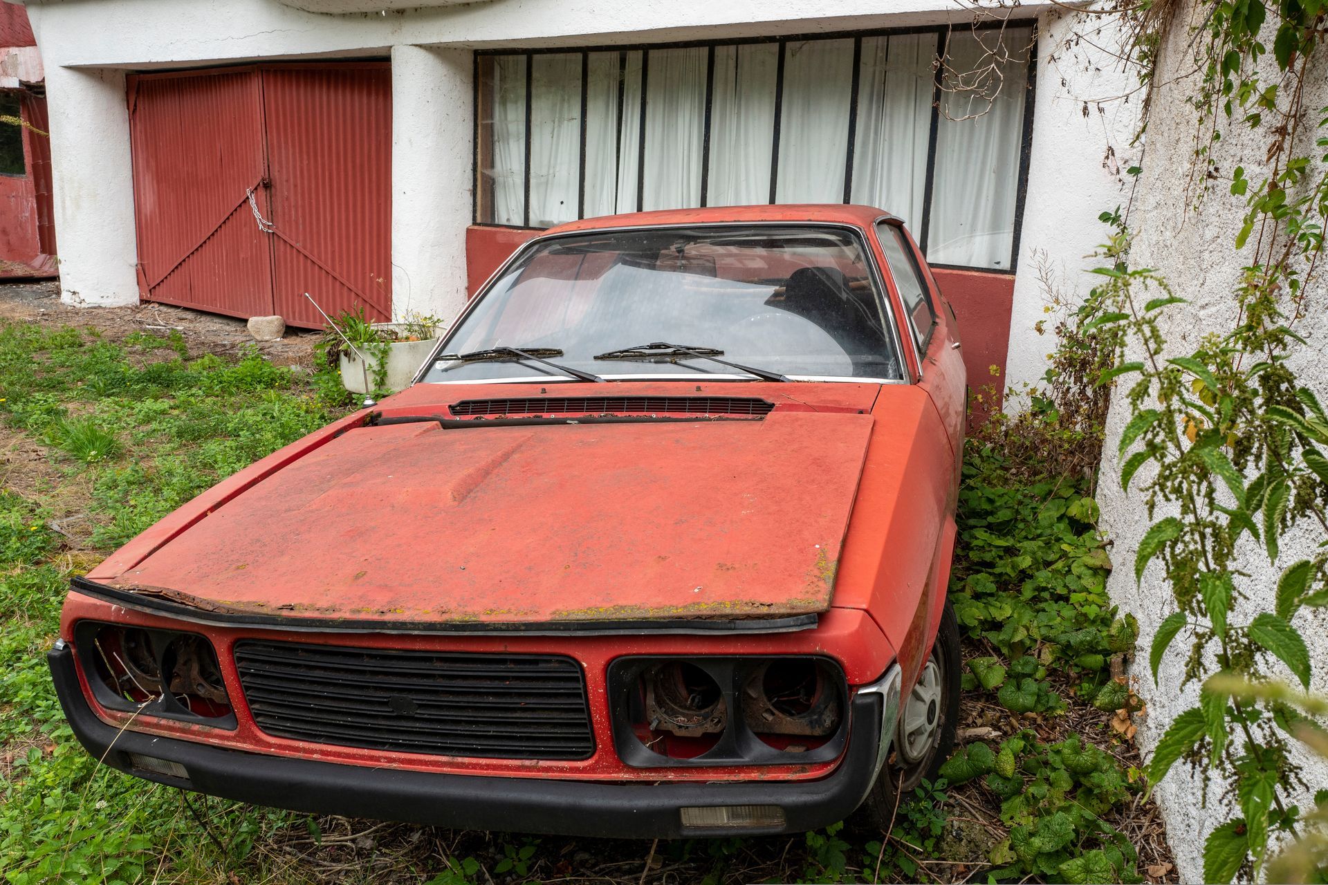 Voiture ancienne rouge, très abîmée par les intempéries, garée près d'un bâtiment aux portes rouges envahi par la végétation.